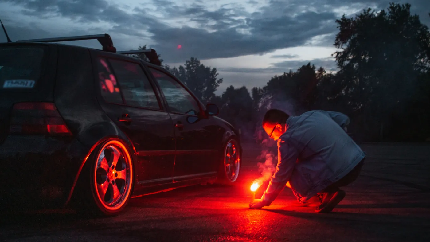 A driver crouched beside his stopped car at dusk, lighting a red road flare